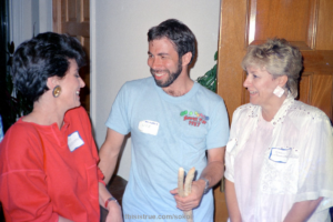 A smiling Woz, dressed in a blue T-shirt with the described stick-on nametag speaking with two ladies inside a house.