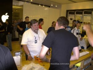 Woz, like other people standing in line wearing a nametag held with a which string around his neck (his says "Woz" of course!) standing at the counter while an employee works on ringing up his purchase. Woz is holding his wallet in his hand, with a credit card visible on top of it.